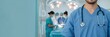 © CoralFox - A close-up of a male doctor with a stethoscope against a medical team performing surgery in an operating room.  Concept of human medical health. Banner with copy space area.