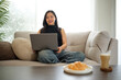 © wattana - Confident young woman smiling at the camera while working on a laptop from her kitchen table with coffee and pastry on the side.