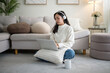 © wattana - Young Asian woman wearing headphones, working on a laptop while sitting cross-legged on the floor of a modern living room.