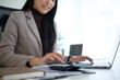 © wattana - Close-up of an Asian businesswoman using a calculator while working on financial documents and her laptop.