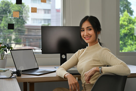 Portrait of confident Asian female graphic designer at work, smiling while sitting at her desk in a contemporary workspace. Creative career and modern technology concept