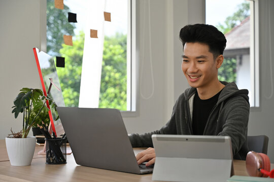 Happy Young Asian male graphic designer focusing on digital projects with multiple devices