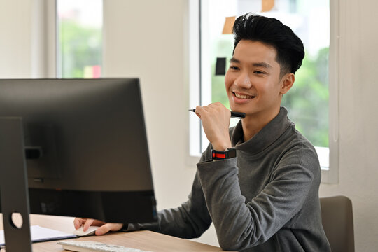 Young Asian graphic artist smiling while working on a desktop computer, illustrating creativity and inspiration in a modern workspace