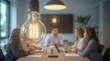 © Ada - Young creative business people meeting at office, brightly lit Edison-style light bulb hangs prominently in the foreground, casting a warm glow over a blurred group of people gathered around a table