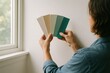 © Татьяна Евдокимова - Person holding paint swatches against a wall near a window, exploring color options for home interior design and renovation projects