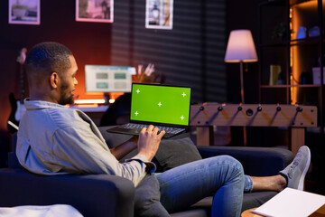Poster - African american young guy using device with green screen from his sofa, enjoys the comfort of his modern cozy home. Casual relaxed person working in the living room, multitasking.