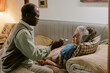 © AnnaStills - Senior Black man feeding senior Caucasian woman lying on couch, both showing signs of aging, man gently holding spoon near woman's mouth, intimate caregiving moment in modest home