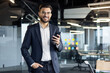 © Liubomir - A smiling businessman in a suit holds a phone in a modern office setting. He looks confident and professional.