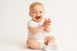 © OlenaSpark - Joyful happy caucasian toddler in white onesie posing in studio. The child smiles brightly while sitting on floor and baby clapping hands