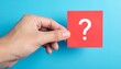 © photostockatinat - A hand holding a red card with a white question mark, set against a blue background. The image poses a query, prompting thought