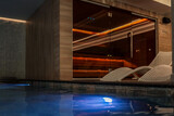 Cozy indoor pool area with modern white lounge chairs and warm-lit sauna behind glass wall. Elegant spa ambiance with wooden textures and relaxing atmosphere for wellness and rest