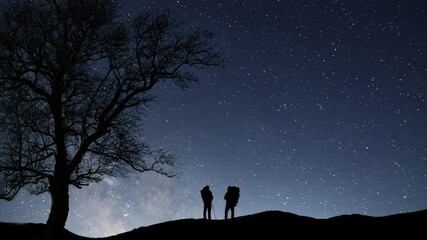 Wall Mural - Two hikers  silhouette with backpack  stands on the hill on the  Milky Way galaxy  background. Night tight landscape starry sky.