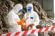 © BESTIMAGE - Two workers in full protective hazmat suits with blue goggles and orange gloves examining a sample or specimen at a hazardous waste site with rocky background and safety barrier