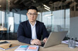 © Tetiana - Portrait of a serious young Asian businessman sitting in the office at his desk, working on a laptop and looking at the camera