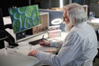 © Seventyfour - Senior Caucasian man with long grey hair and beard working at laboratory desk analyzing scientific data on computer monitor displaying cell structures, writing notes on paper