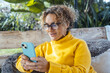 © simona - Attractive woman in yellow sweater uses cell phone and relaxes alone in the garden sitting on a sofa. Portrait of serene female writing on cell phone. Mature young people relaxing