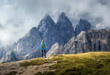 © den-belitsky - Sporty woman standing on grassy slope and admiring dramatic mountain range in clouds in Dolomites, Italy. Summer travel in Alps. Trekking and hiking. Girl with backpack in alpine mountains. Landscape