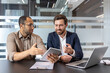 © Liubomir - Two businessmen in a modern office setting collaborate, reviewing data on a tablet and laptop, during a discussion about business.