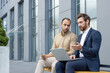 © Liubomir - Two business professionals collaborating outdoors near a modern building, using a laptop and a tablet.