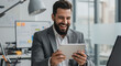 © Luiz - A bearded man in a suit laughing while holding a tablet in an office setting with a computer nearby