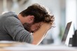 © ttonaorh - Unhappy businessman suffering from neck pain in the office, laptop on the desk, blurred background. Distressed man with his head between his hands and face down at the work table.