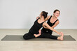 © TRAVELARIUM - Female yoga instructor guides adult woman in seated stretch on yoga mat. Minimalist studios neutral tones highlight focus and calm. Both wear black outfits, emphasizing form and balance