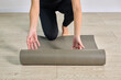 © TRAVELARIUM - Young woman in black leggings setting foam roller on light wooden floor, indoor exercise setup. Scene suggests focus on fitness, relaxation, and self-care