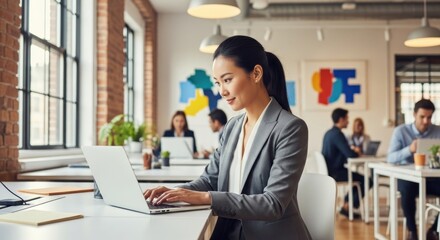 Woman in a gray suit working on a laptop in a modern office with other people in the background
