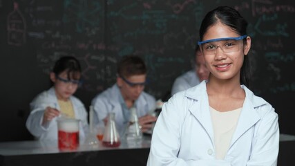 Poster - high school girl looking at camera while people doing experiment at laboratory. Young cute student pose at camera while standing blackboard with chemical theory with blurring background. Edification
