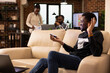 © DC Studio - Smiling businessman sits on couch in brick wall office, interacting with glowing 3D hologram. African American male employee holding holographic handheld device and waving during virtual meeting.