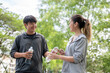 © bongkarn - Asian man in sportswear holding water bottles and giving to a woman with phone at park's courtyard.