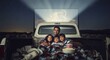 © Leo - A cozy family cuddled together with blankets and popcorn in the back of a pickup truck at a drive-in movie theater at night.