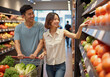 © Akaka Studio - Waist up portrait of a smiling young Asian couple with a shopping cart choosing fruit and vegetables in a supermarket.