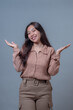 © Saddha Visual - Portrait of a happy young Indonesian woman with long hair, smiling while raising both hands in a questioning or presenting gesture, wearing brown casual outfit, studio shot.