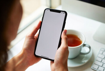 Poster - Female hands holding smartphone with blank screen, near a tea mug and keyboard. Smartphone in hand is mockup for easy customization.