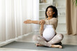 © Anastasiya - Black pregnant woman stretching arms while seated on yoga mat. Joyful moment of prenatal fitness and flexibility, promoting wellness and healthy lifestyle at home, enjoying meditation
