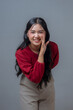 © Saddha Visual - Indonesian woman smiling brightly while making a whispering gesture with her hand near her mouth. Young Asian female in red top posing playfully against gray background.