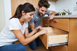 © Alex & M Studio - Happy young couple working together as a team to assemble a new wooden kitchen drawer, smiling while doing a DIY home improvement project in their modern apartment