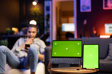 Poster - Young relaxed man with headset having a green screen in cozy living room, stylish space decorated with furniture and neon lights. Casual guy relaxing on a bean bag at home, multitasking.