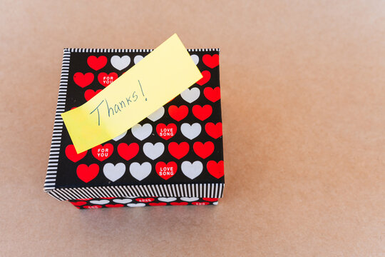 Thank you note that says thank you handwritten with blue pen with a background of a gift box with red and white hearts