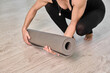 © TRAVELARIUM - Young woman rolling gray yoga mat on light wooden floor. Scene evokes calm and preparation in minimalist indoor setting, viewed from above