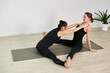 © TRAVELARIUM - Young adult women engaging in partner yoga on wooden floor, one assisting other in stretch. Setting is calm, with minimal decor and natural lighting, evoking tranquil mood
