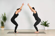 © TRAVELARIUM - Two adult women practice yoga together in minimalist room. Dressed in black activewear, they are positioned on yoga mat, engaging in focused physical exercise