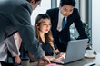 © AU USAnakul - Female office worker typing on laptop with serious expression as she discusses project with team in corporate environment.