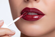 © Marut - Glossy dark red lip tint being applied with applicator wand on smooth skin lips, creating shiny and elegant makeup look with rich color and precise detail