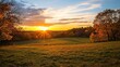 © Phonphysith - A vibrant autumn sunset casts golden light over a grassy field bordered by colorful fall trees under a partly cloudy sky.