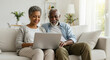 © Artem Zatsepilin - old african american couple sitting on sofa happily using laptop in living room. cozy home, technology, relaxation, online shopping, remote work. sales and discounts. senior activity, active lifestyle
