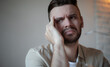 © My Ocean studio - Young man in casual shirt looking stressed and tired, touching his face with concern, sitting at home, natural light, concept of headache, burnout, or overthinking.