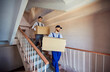 © Studio Romantic - Men movers carrying big packed cardboard boxes with belongings, posing on stairs in entrance, help client move out of apartment, wearing uniform, working in removal service, being good team