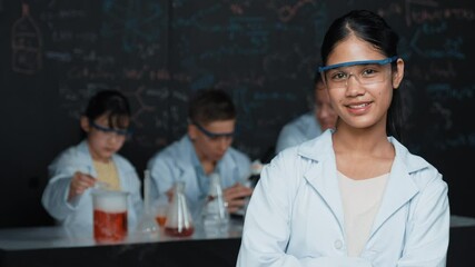 Poster - high school girl looking at camera while people doing experiment at laboratory. Young cute student pose at camera while standing blackboard with chemical theory with blurring background. Edification